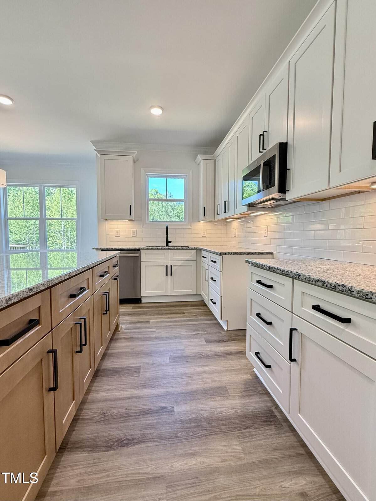 Modern kitchen with white cabinets, granite countertops, stainless steel appliances, and black hardware. Large window lets in natural light. Subway tile backsplash and light wood flooring are visible.