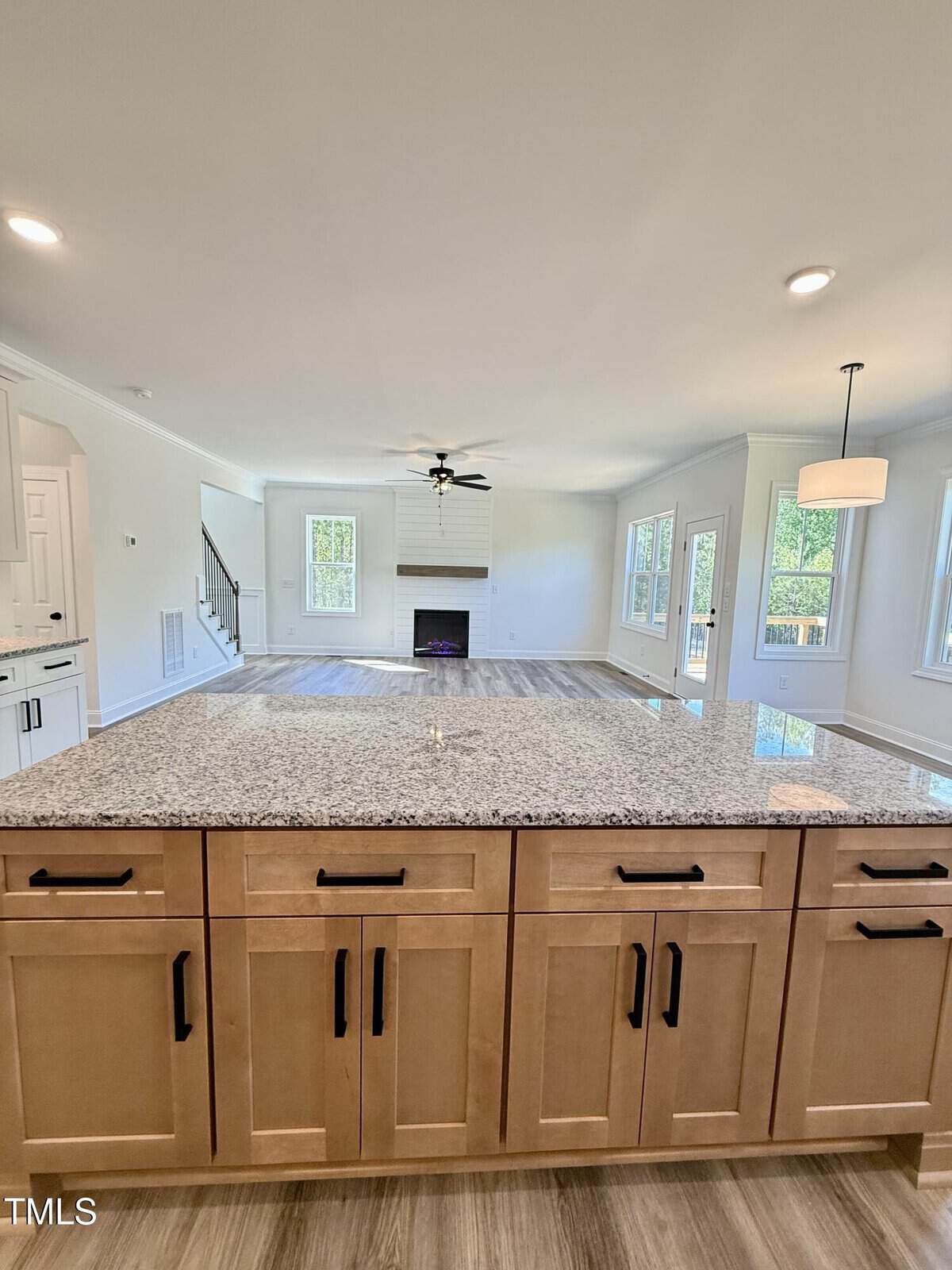 View from a kitchen island with light wood cabinets and granite countertop, looking into an open living area with a fireplace, windows, and a ceiling fan.