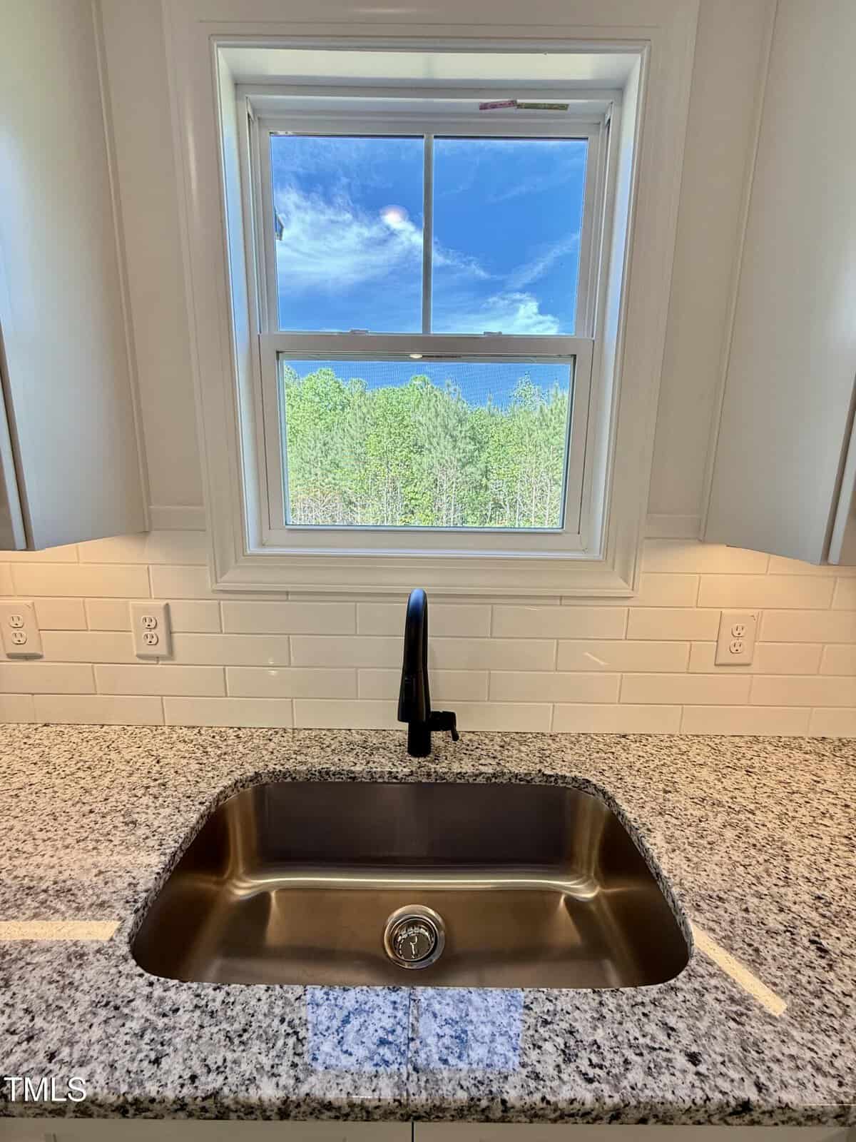 A stainless steel kitchen sink with a black faucet is set in a granite countertop beneath a window, showing a view of trees and a blue sky. White subway tile backsplash and electrical outlets are visible.