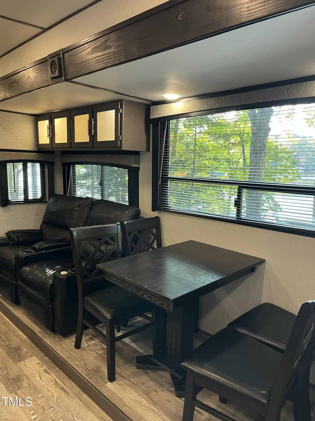 A modern RV interior featuring a black dining table with chairs, a black leather sofa, large windows with blinds, wood flooring, and overhead cabinets. Trees can be seen outside through the windows.