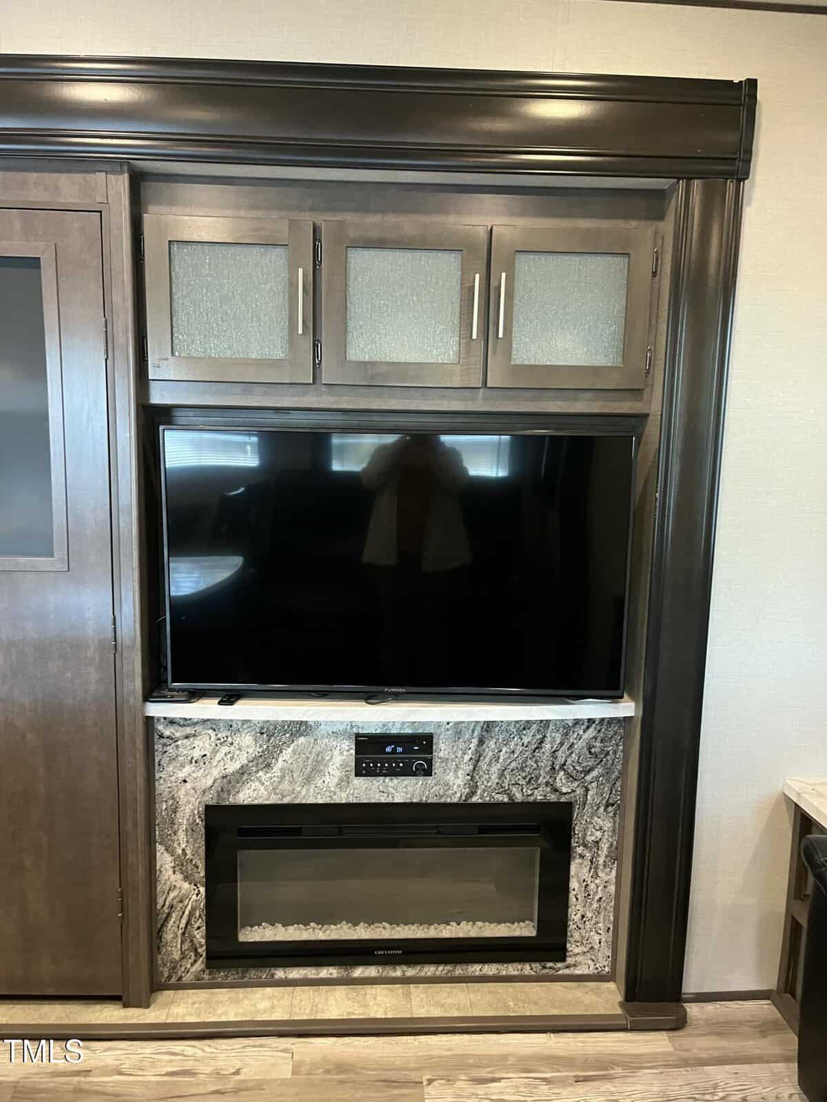 Wood entertainment center with three cabinets above a flat-screen TV. Below the TV is a marble-patterned electric fireplace and control panel, with part of a door and countertop visible on the sides.