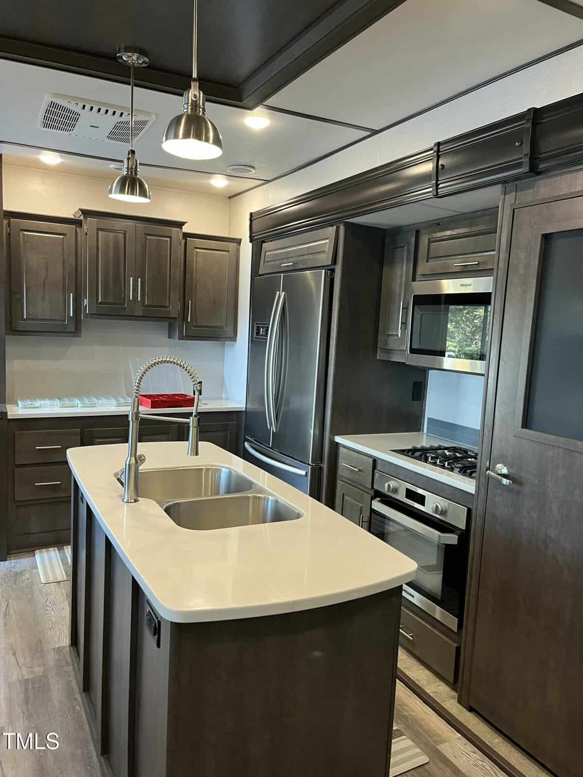 Modern kitchen with dark wood cabinets, stainless steel appliances, a gas range, microwave, double-door fridge, island with sink and pull-down faucet, pendant lights, and light countertops.