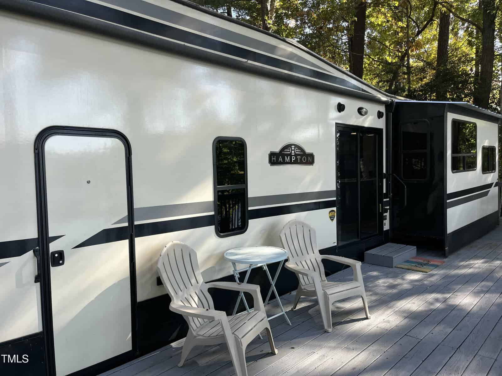 A white and black RV with a "Hampton" logo is parked beside a gray wooden deck. Two white Adirondack chairs and a small round table are on the deck. Trees with green leaves are visible in the background.