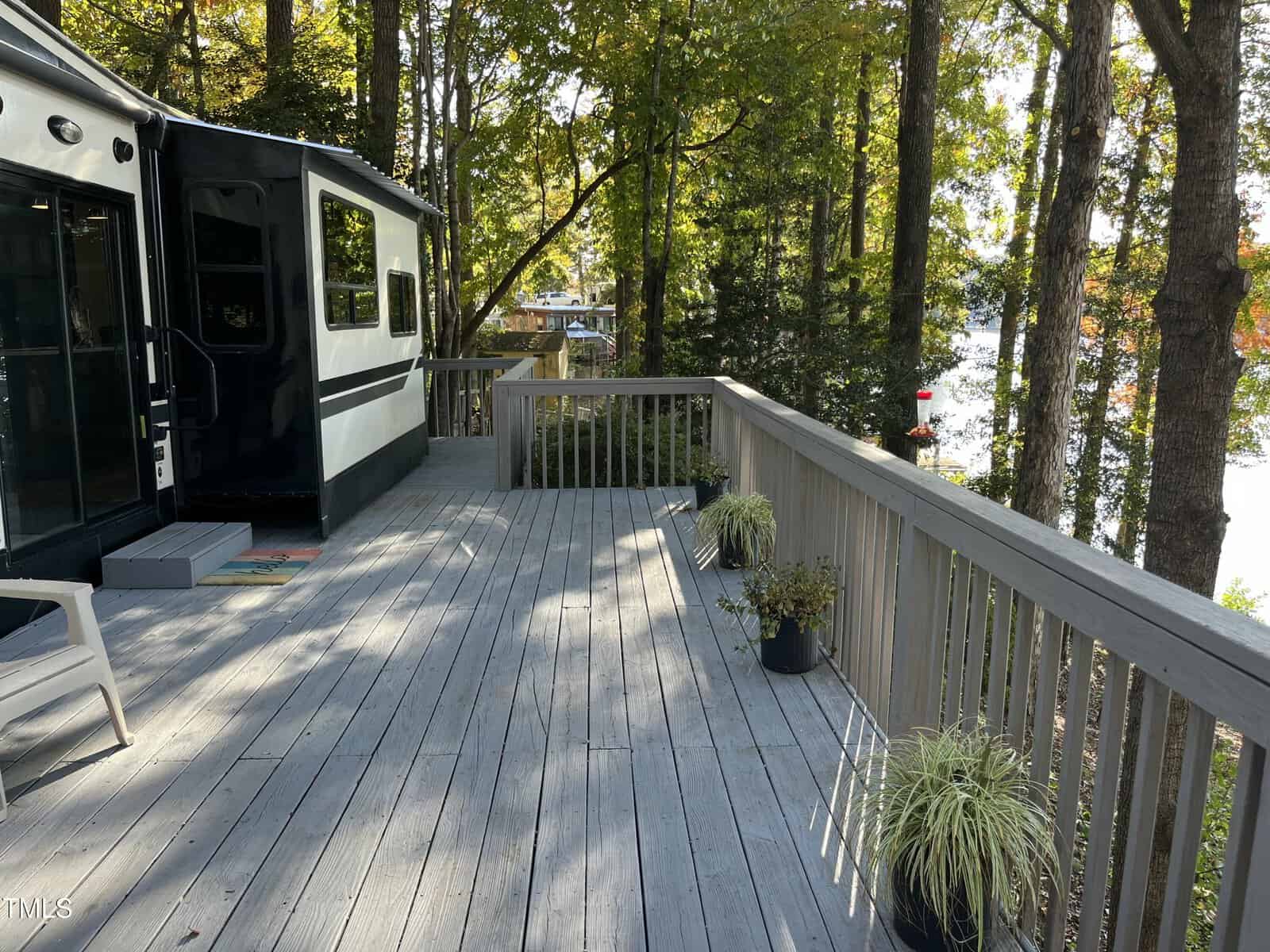 A wooden deck with potted plants and a chair is attached to a black and white trailer, surrounded by trees. A lake is visible through the trees in the background.