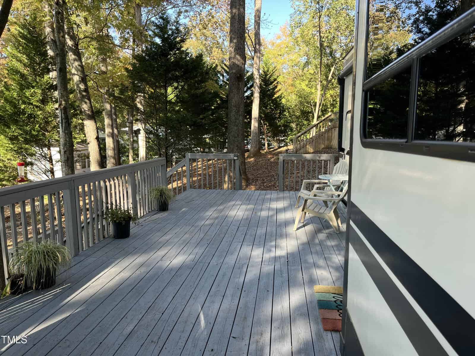 A spacious wooden deck with gray railings, two white chairs, potted plants, and a doormat. Trees and a fence are in the background; part of a vehicle is visible on the right.