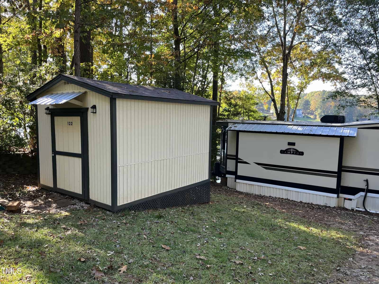 A small beige and black shed with the number 122 on the door sits next to a white travel trailer in a grassy, wooded area with trees and fallen leaves. Sunlight filters through the trees.