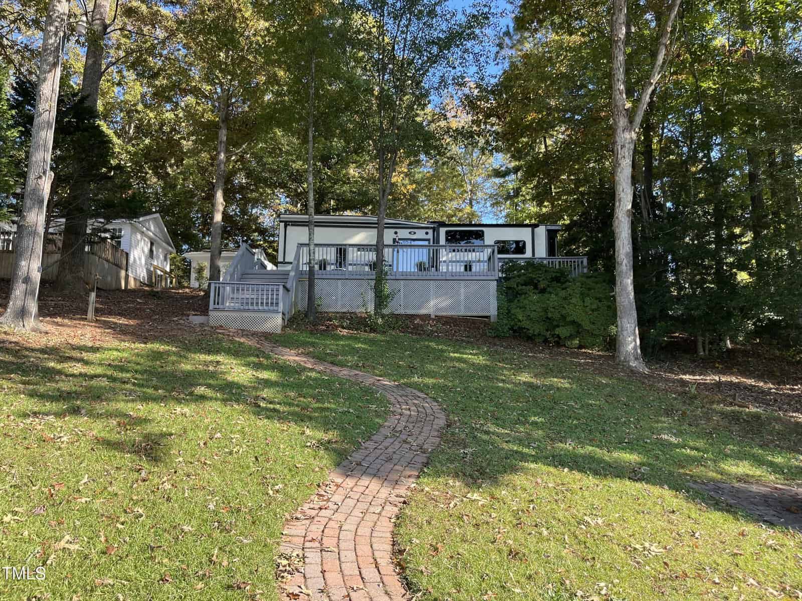 A brick pathway leads uphill through a grassy lawn to a raised wooden deck and a white RV or mobile home, surrounded by tall trees with green and autumn-colored leaves.