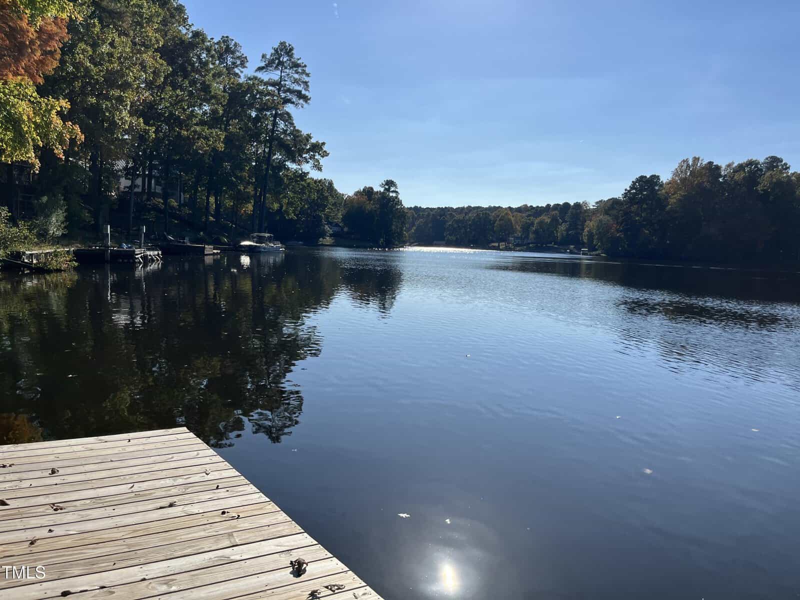 A wooden dock extends over the edge of a calm lake surrounded by trees with green and autumn-colored leaves on a clear, sunny day. The sky is blue and the water reflects the trees and sky.