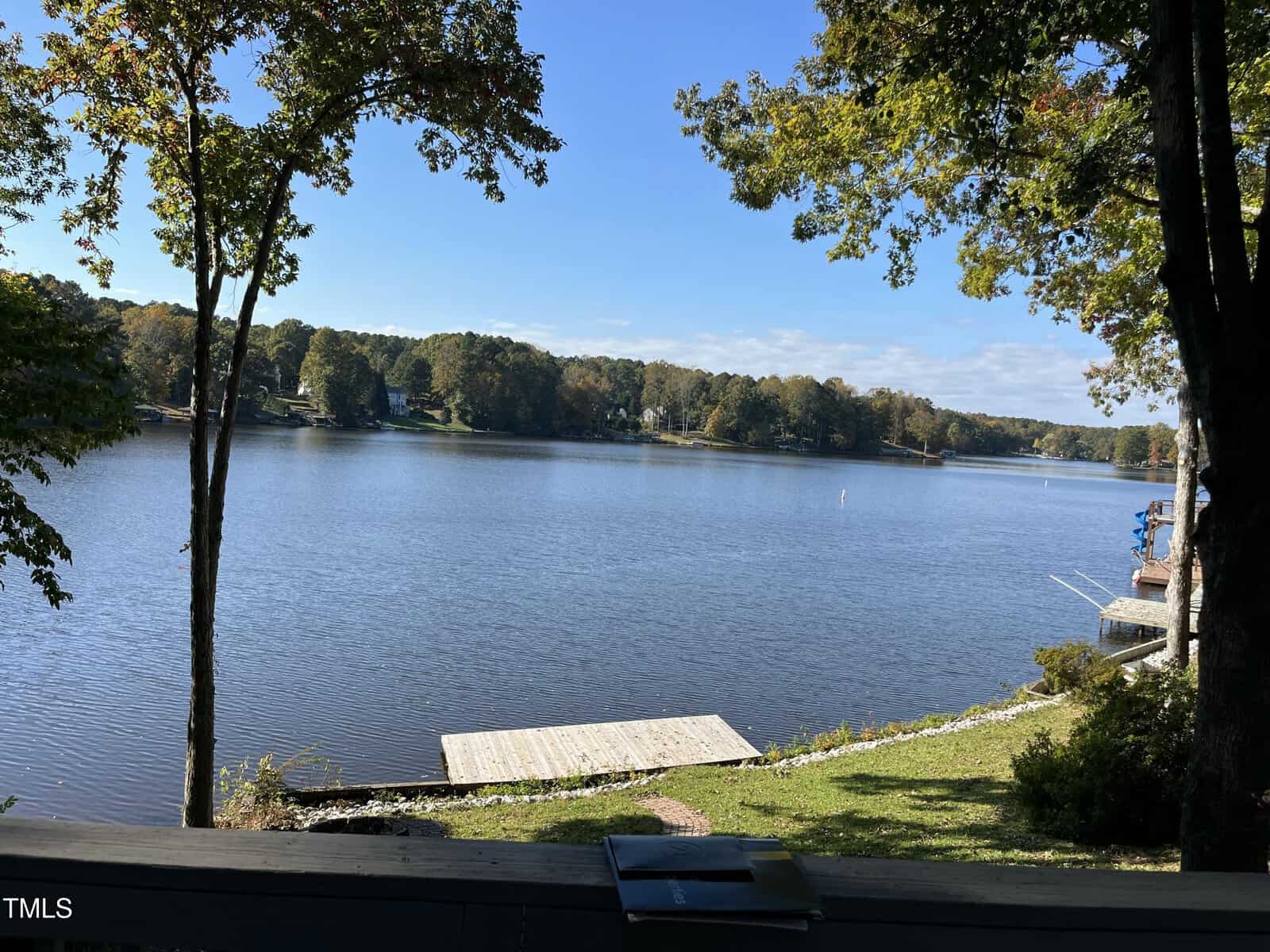 View of a calm lake with a wooden dock extending from the shore, surrounded by trees with green foliage. Houses are visible on the far side of the lake under a clear blue sky.