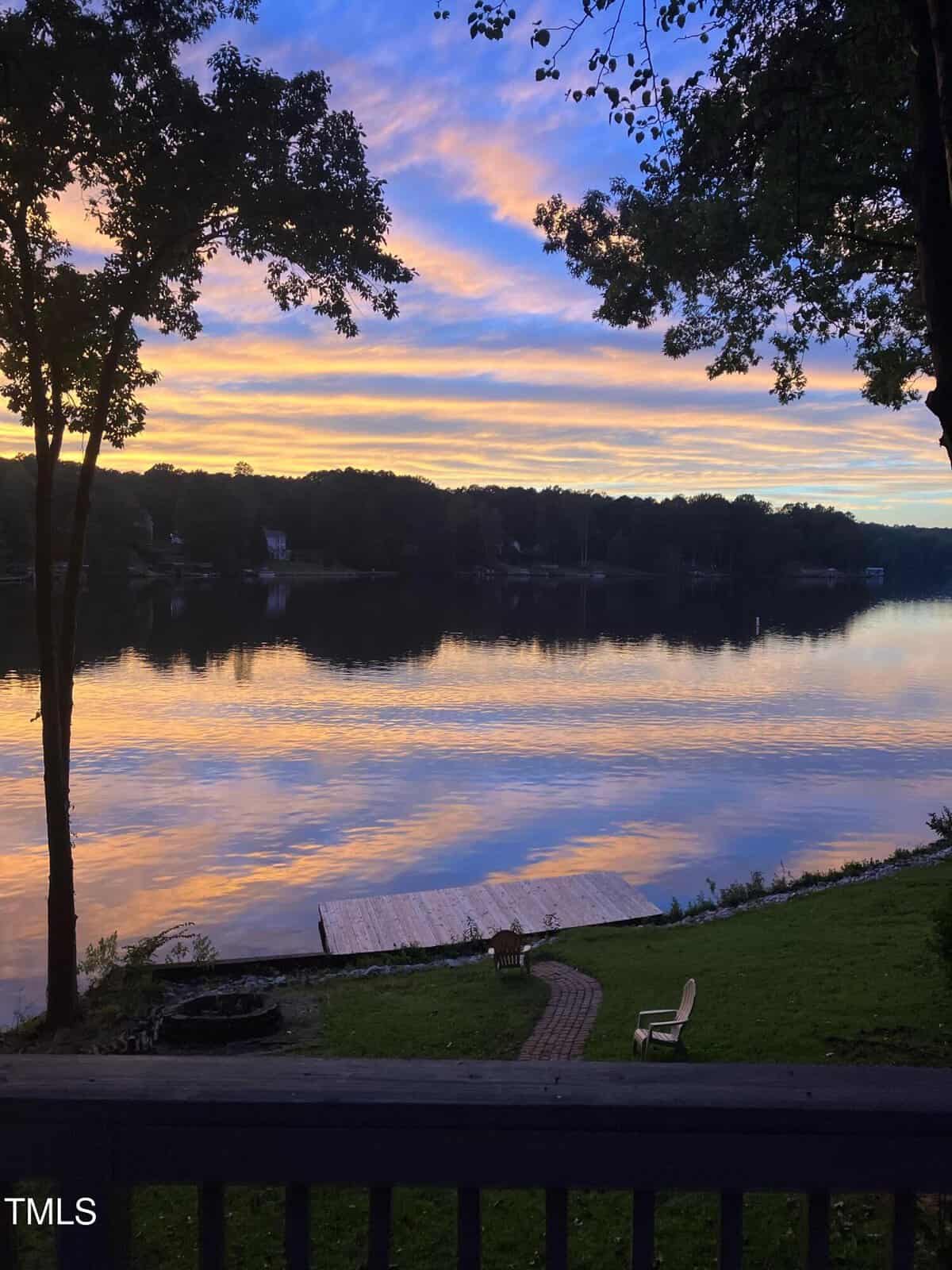 A calm lake at sunset with reflections of trees and clouds on the water, a dock extending into the lake, grassy shore, a path, and several outdoor chairs in the foreground.