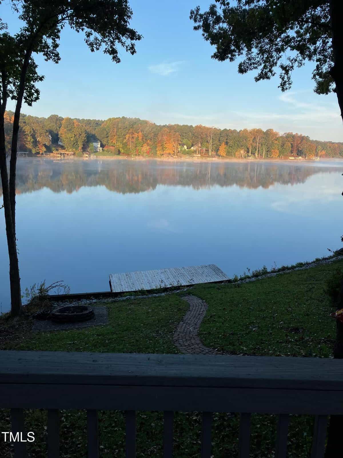 View of a calm lake with a wooden dock, surrounded by trees with autumn foliage. The scene is seen from a porch with a railing, and a fire pit is visible on the grassy lawn leading down to the water.
