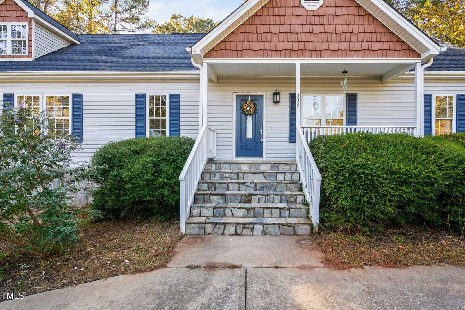 A house with light-colored siding, blue shutters, a blue front door with a wreath, white railings, and a stone staircase leading to the entrance; bushes and a concrete walkway are in front.