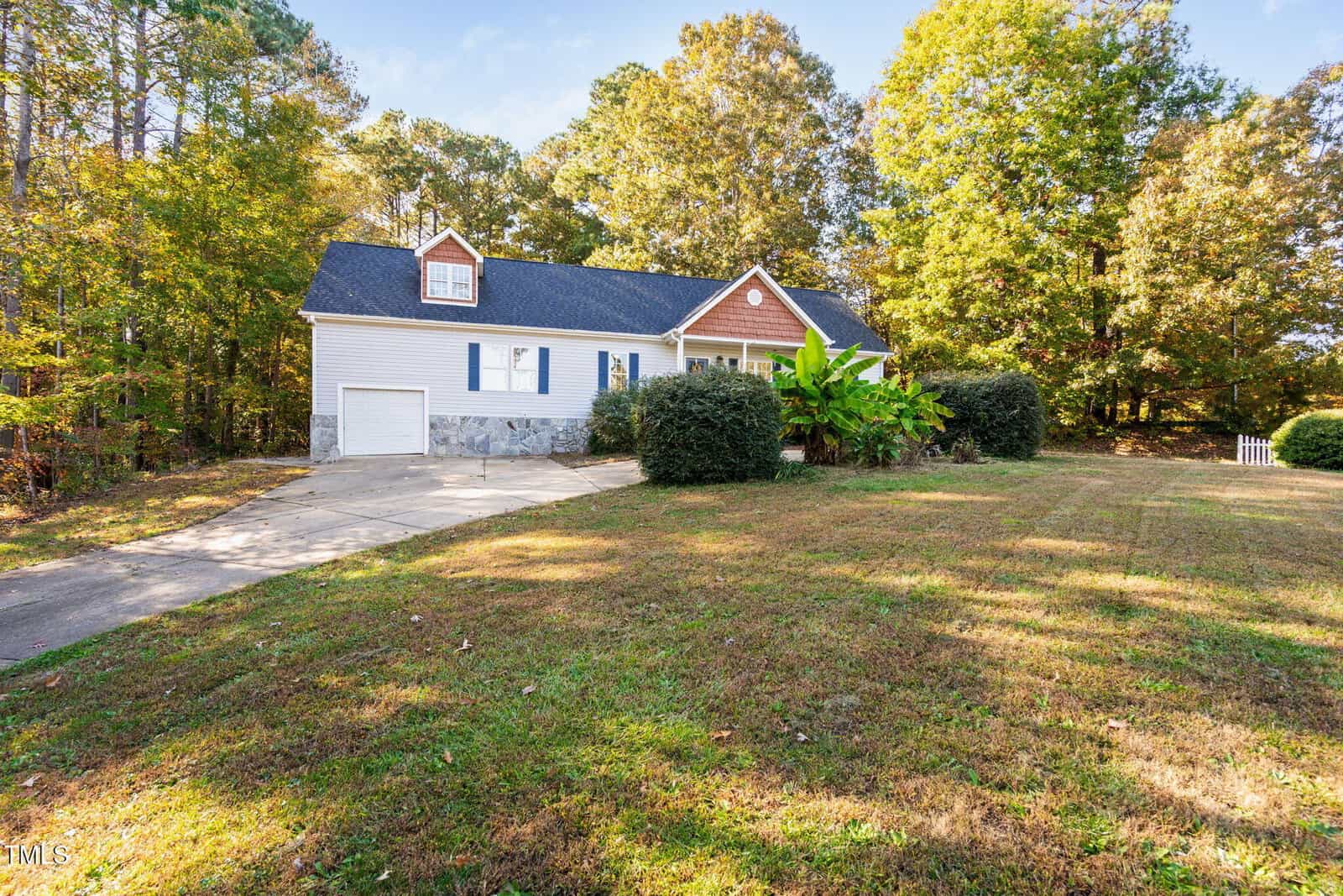 A single-story house with white siding, a dark roof, and a stone foundation sits beside a driveway. The front yard is grassy with bushes and trees surrounding the property. The sky is clear and blue.