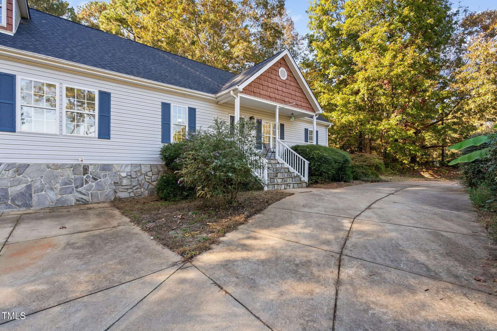 Single-story house with white siding, blue shutters, and stone foundation. A covered front porch with steps leads to the entrance. Curved concrete driveway and bushes are in front, with trees and foliage in the background.