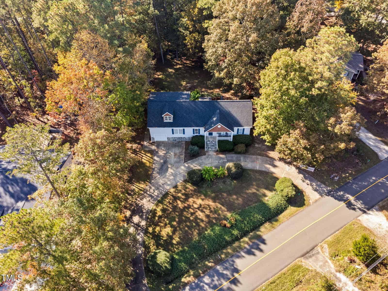 Aerial view of a single-story house with a dark roof and light-colored exterior, surrounded by trees and greenery, featuring a curved driveway and located next to a two-lane road.