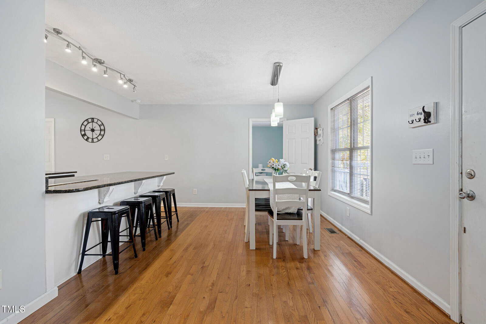 A dining area with a white table and four chairs sits beside large windows; a breakfast bar with four black stools is on the left, and the room features light blue walls and hardwood floors.