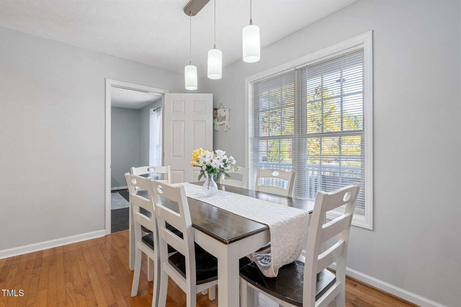 A dining area with a white table and six chairs, a light fixture with three pendant lights above, a vase with flowers on the table, wood flooring, and a large window with blinds letting in natural light.