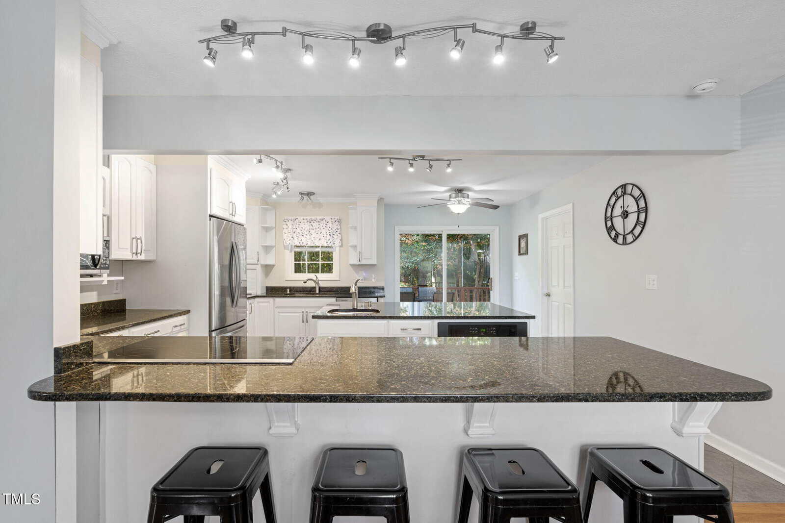 Modern kitchen with white cabinets, stainless steel appliances, black granite countertops, four black stools at an island, track lighting, ceiling fan, large window, and a wall clock. Sliding glass doors lead to a green backyard.
