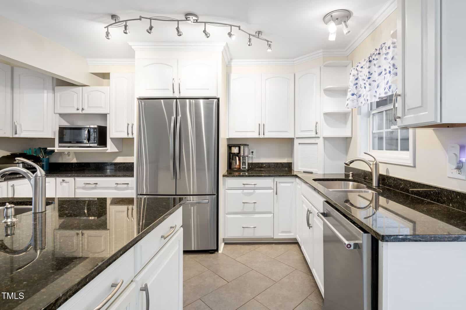 Modern kitchen with white cabinets, stainless steel appliances, black granite countertops, and tiled floor. Track lighting and a window with a patterned valance provide ample light. There is a double-door fridge and built-in microwave.