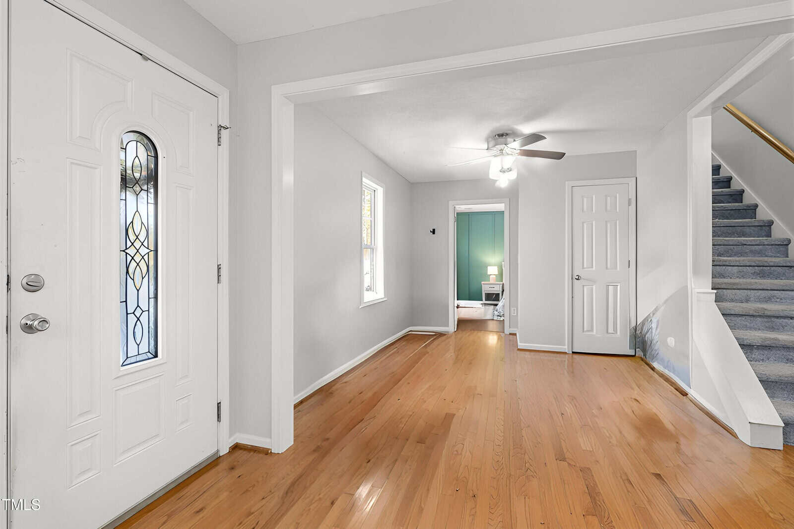 A view of a bright entryway with a white front door, hardwood floors, a ceiling fan, a staircase with gray carpet, and a doorway leading to a room painted in teal.