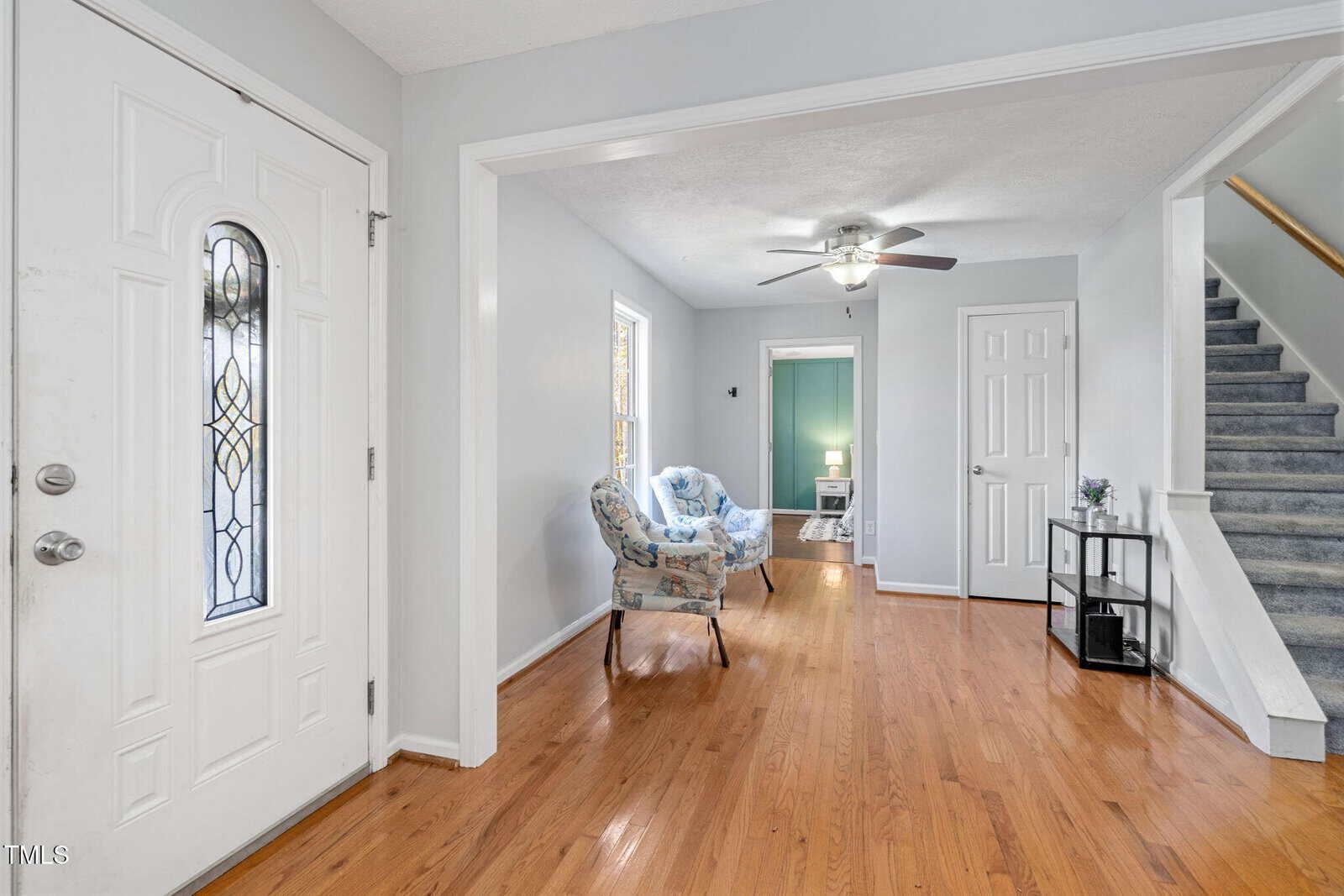 A view of a home's entryway with a white front door, hardwood floors, two floral chairs in a sitting area, a small table with a lamp in a blue room, staircase, and a white door under the stairs.