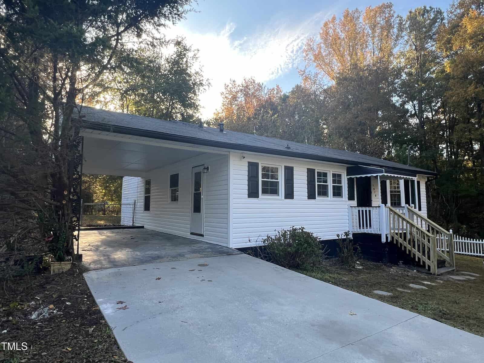 A single-story white house with black shutters, surrounded by trees. It features a covered carport and a small porch with railings at the front entrance. The driveway leads to the carport. The yard is neatly maintained.
