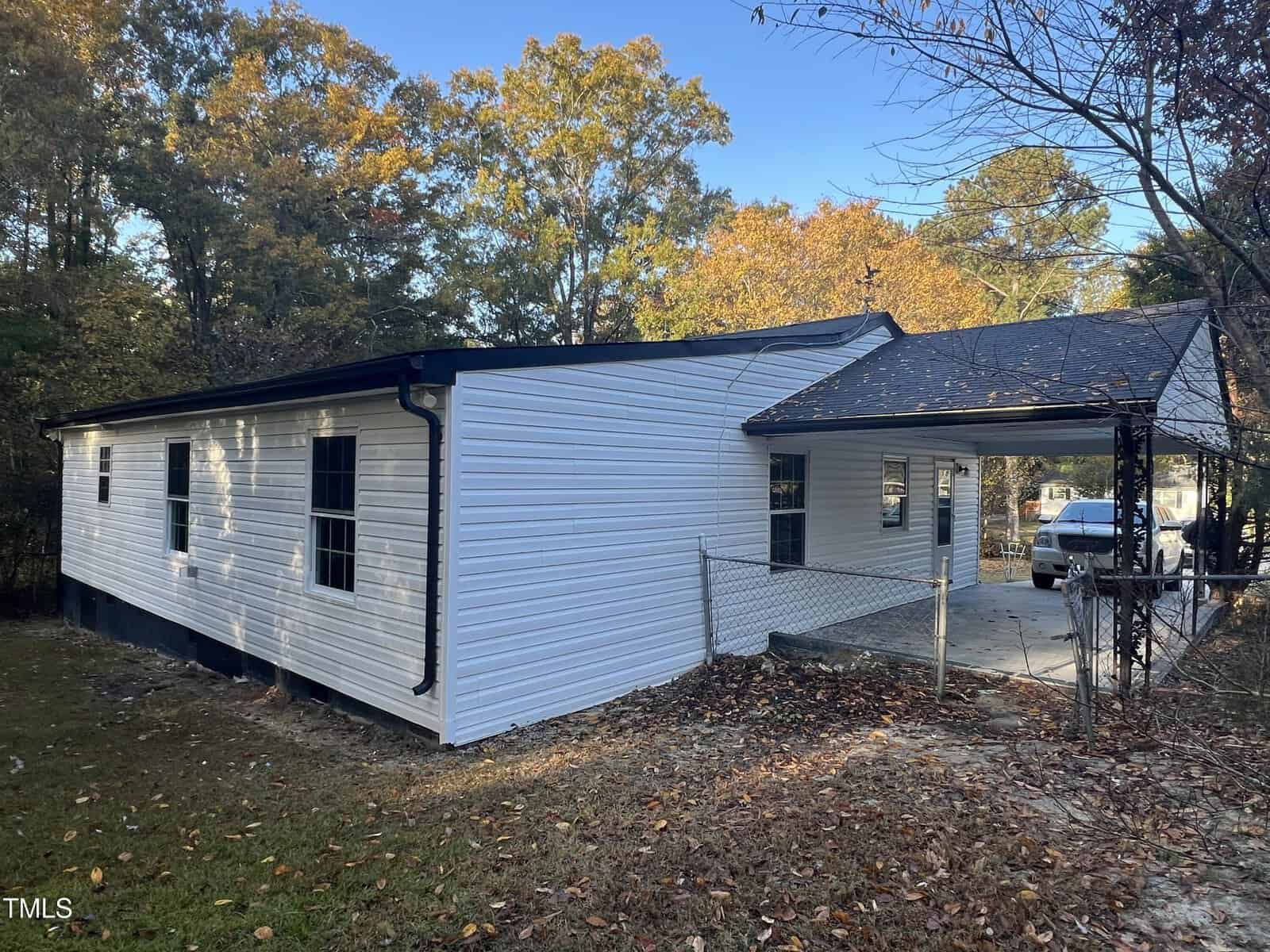 A white, single-story house with a metal roof and attached carport is surrounded by trees. The yard is fenced, and a car is parked in the driveway. Fallen leaves cover parts of the ground, indicating an autumn setting.