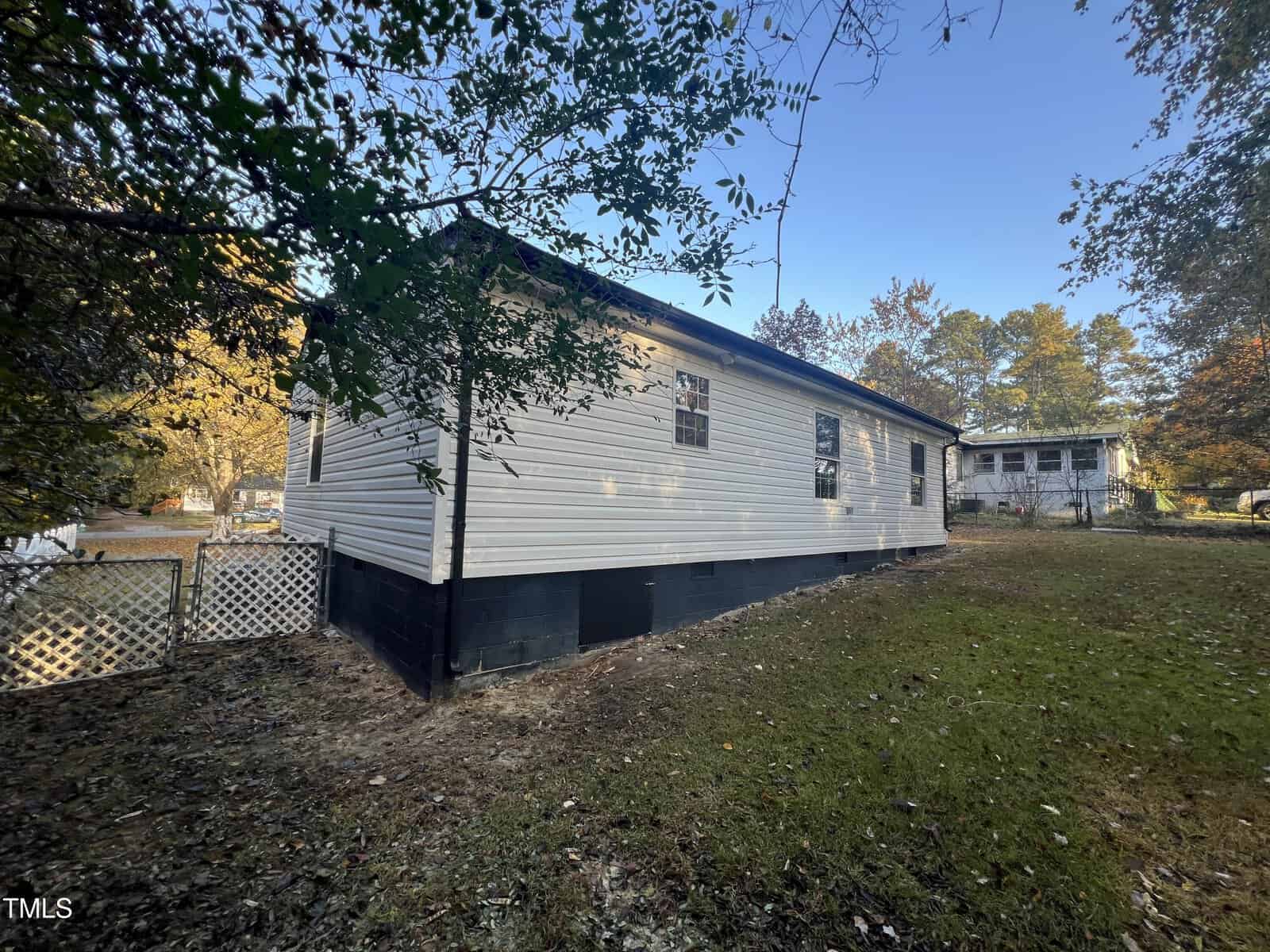 A single-story white mobile home with a dark foundation sits on a grassy lot. A white lattice fence borders one side. Trees with fall foliage are in the background under a clear blue sky.