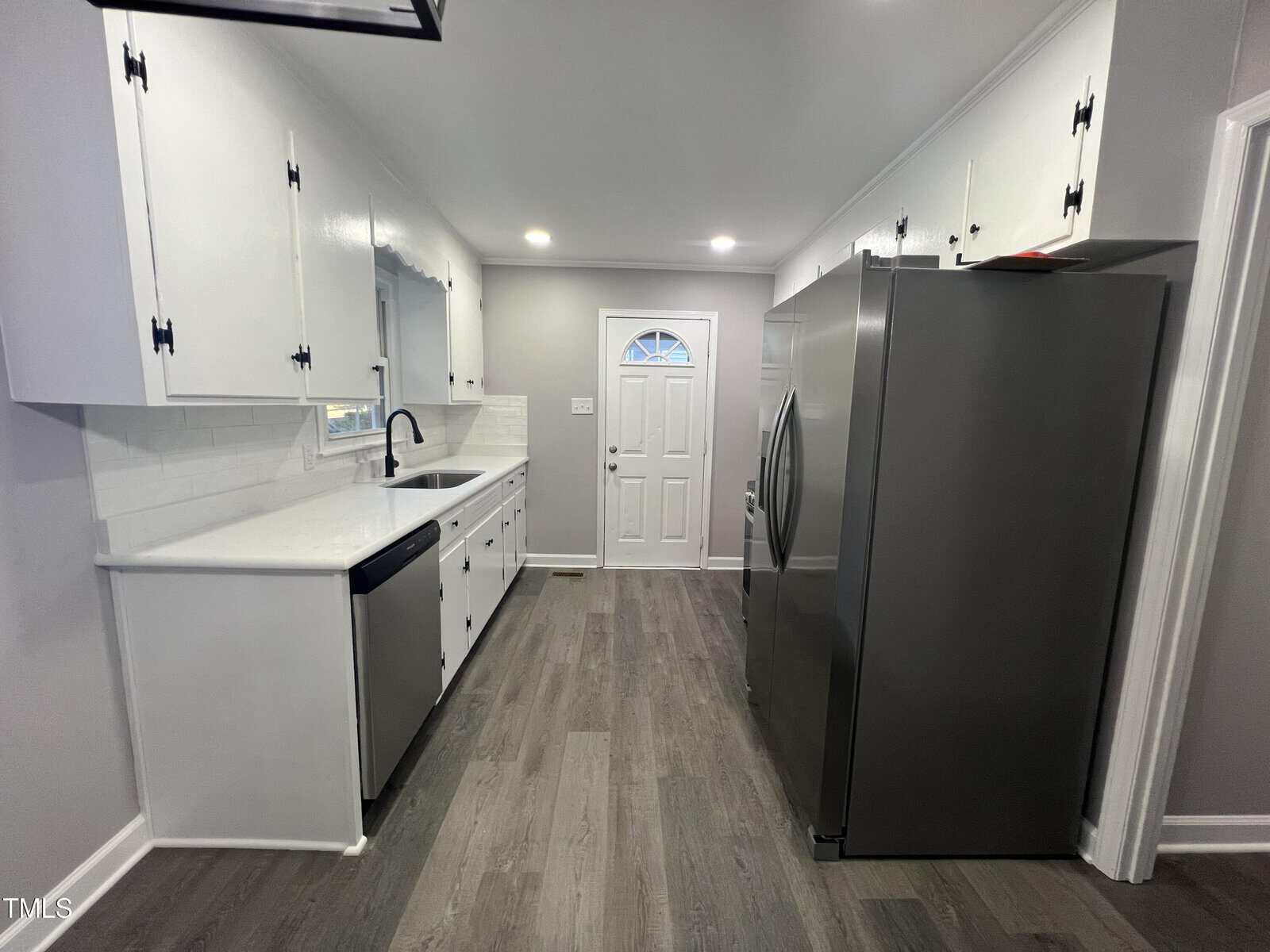 A modern kitchen with white cabinets, a stainless steel sink, and a large silver refrigerator. The floor is gray wood, and there's track lighting on the ceiling. A white door with a decorative window is at the end of the room.