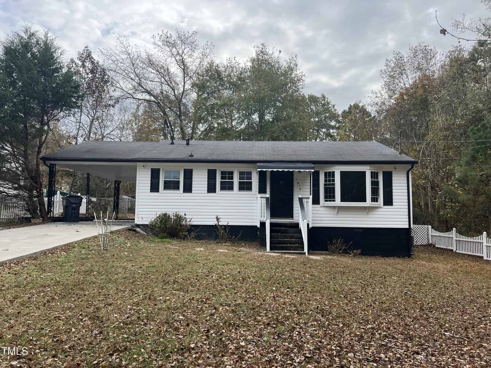 Nestled at 1910 Willow Lane, a small white house with black shutters graces a leaf-covered lawn. It features a carport on the left, steps leading to the front door, and trees standing tall in the background beneath Henderson's cloudy sky.