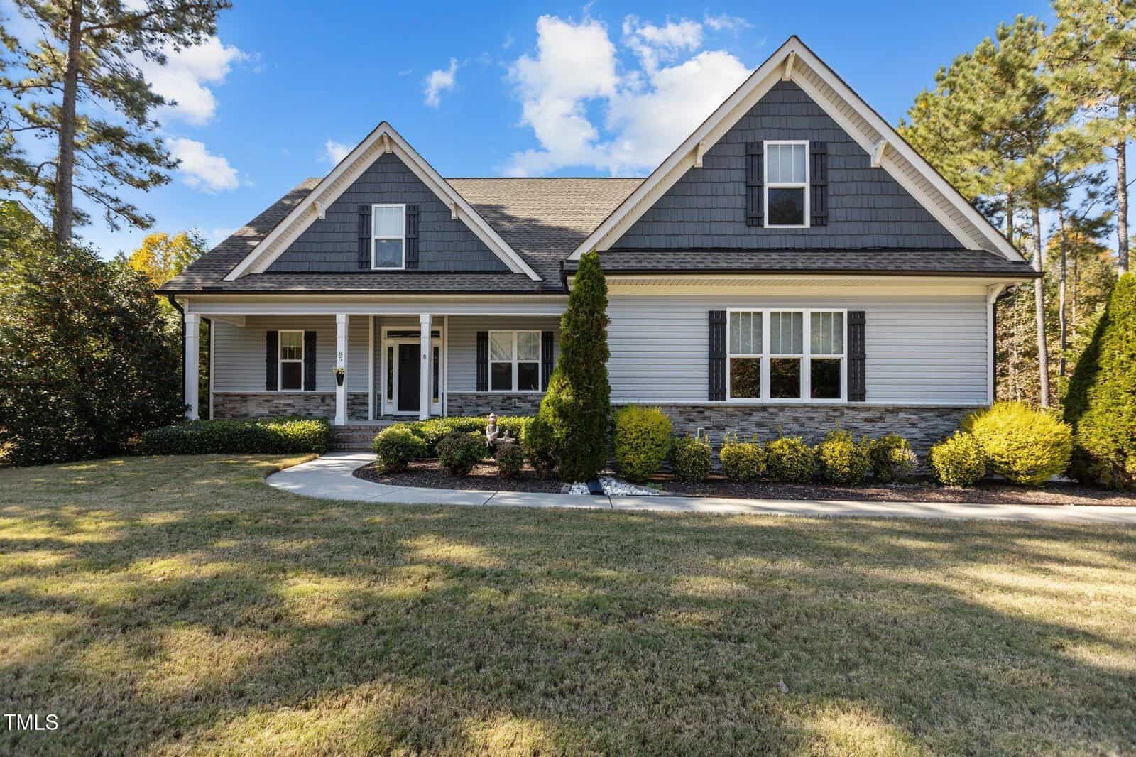 A two-story suburban house with gray siding, dark shingles, stone accents, and a covered front porch, surrounded by neatly trimmed bushes, tall trees, and a well-kept lawn under a blue sky with scattered clouds.