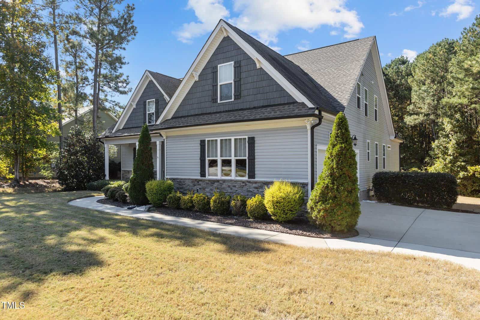 A gray suburban house with dark shingles, white trim, and a stone accent, featuring manicured bushes, a curved walkway, and a concrete driveway surrounded by green trees and a sunlit lawn.