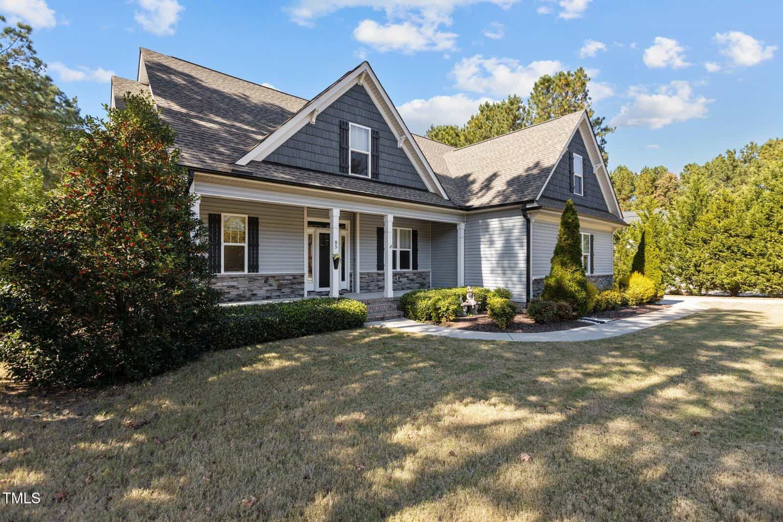 A two-story gray house with white trim, gabled roof, and stone accents, surrounded by trimmed bushes, trees, and a manicured lawn under a blue sky with scattered clouds.