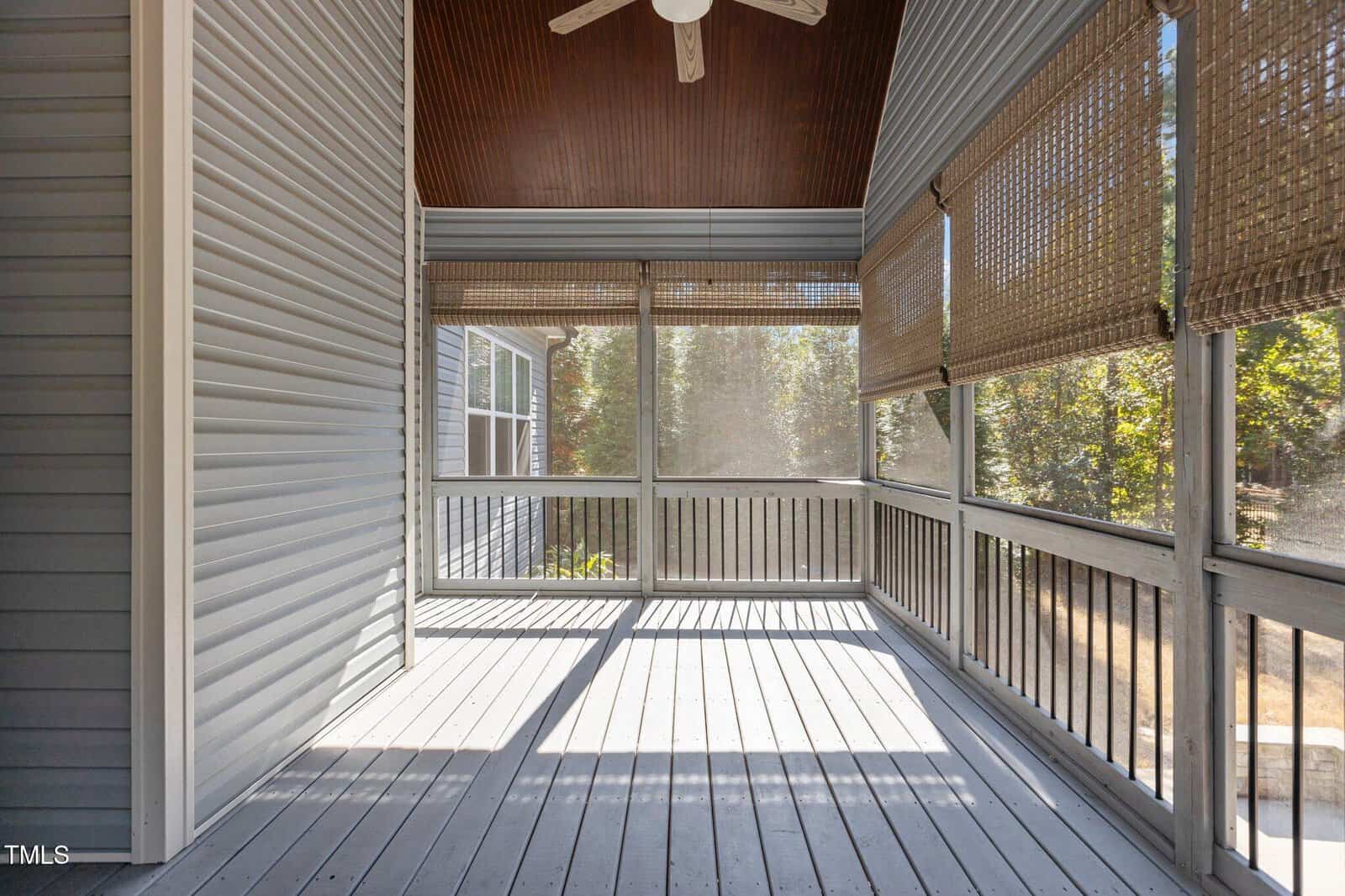 Screened-in porch with gray wooden floor, light gray siding, and a high, wooden-paneled ceiling with a ceiling fan. Bamboo shades cover the screened windows, and sunlight streams in, casting shadows on the floor.