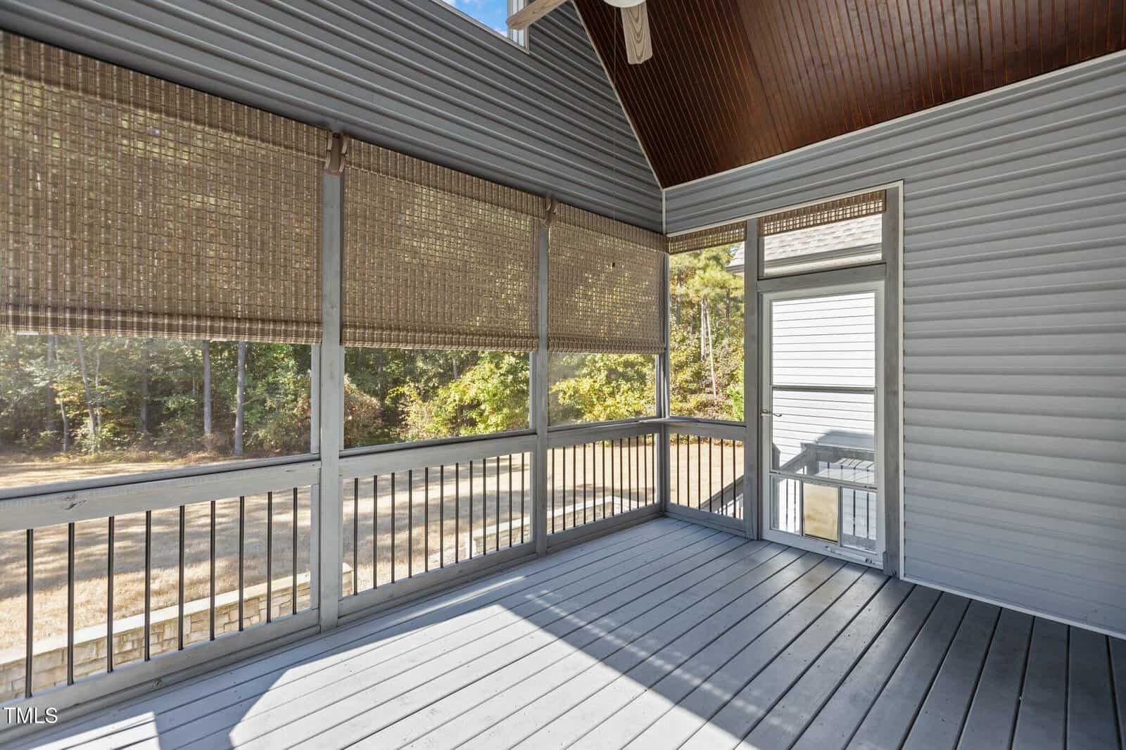 A screened-in porch with gray wooden flooring, light gray siding, bamboo roll-up shades, and a vaulted ceiling with dark wood paneling. Trees and grass are visible outside through the screened windows.