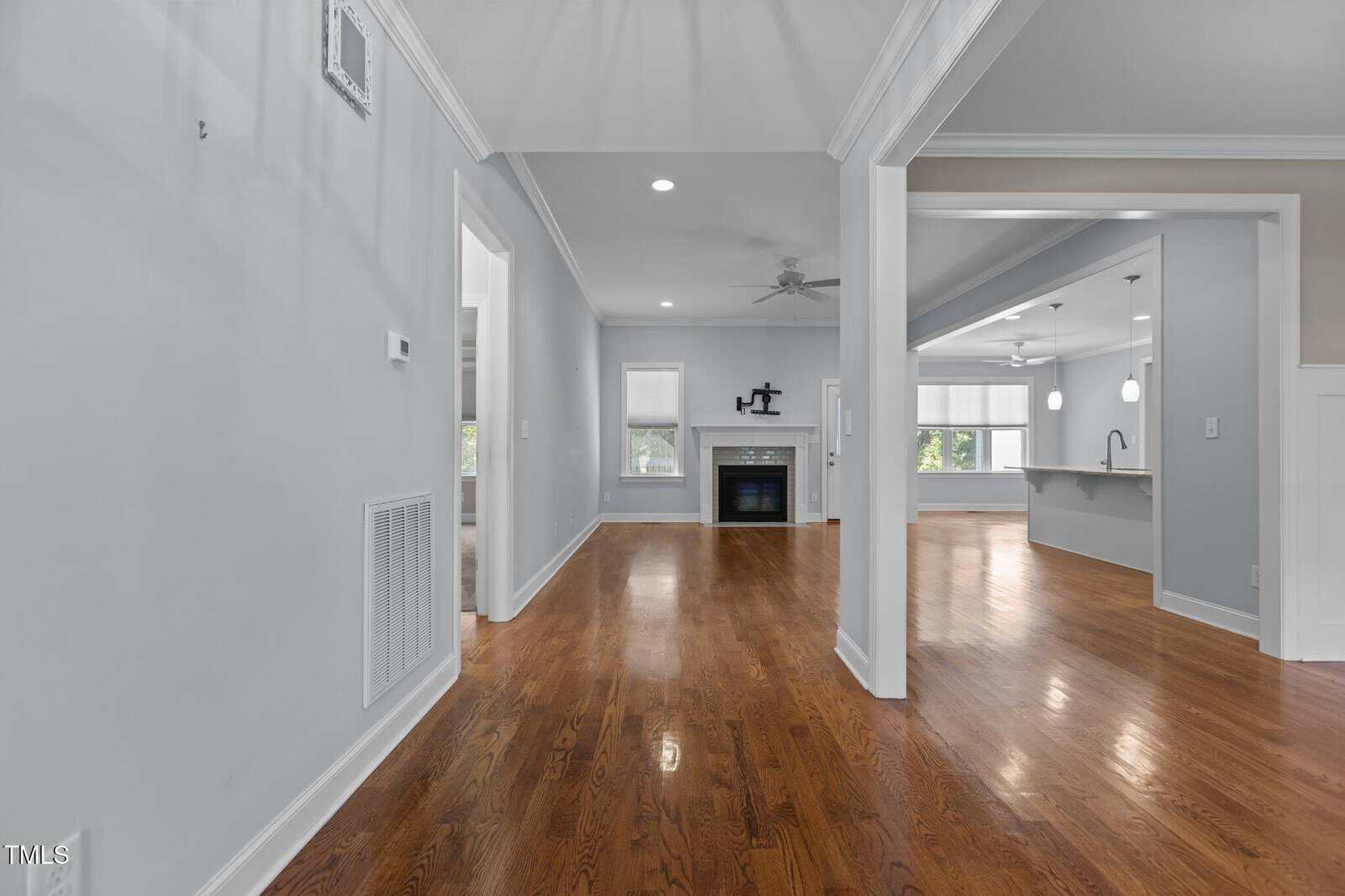 Spacious modern home interior with hardwood floors, light gray walls, and white trim. The hallway leads to a living area with a fireplace, ceiling fan, large windows, and an open kitchen with a breakfast bar.