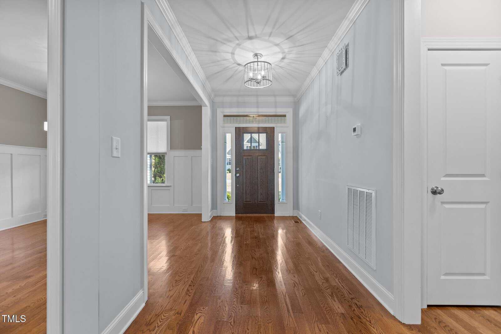 Bright entryway of a house with hardwood floors, white walls, a modern chandelier, and a wooden front door with glass panels. Two rooms are visible on either side of the hallway.