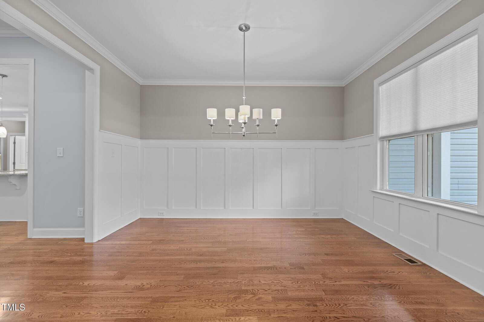 A bright, empty dining room with hardwood floors, white wainscoting, light gray walls, a modern white chandelier, and two large windows letting in natural light.