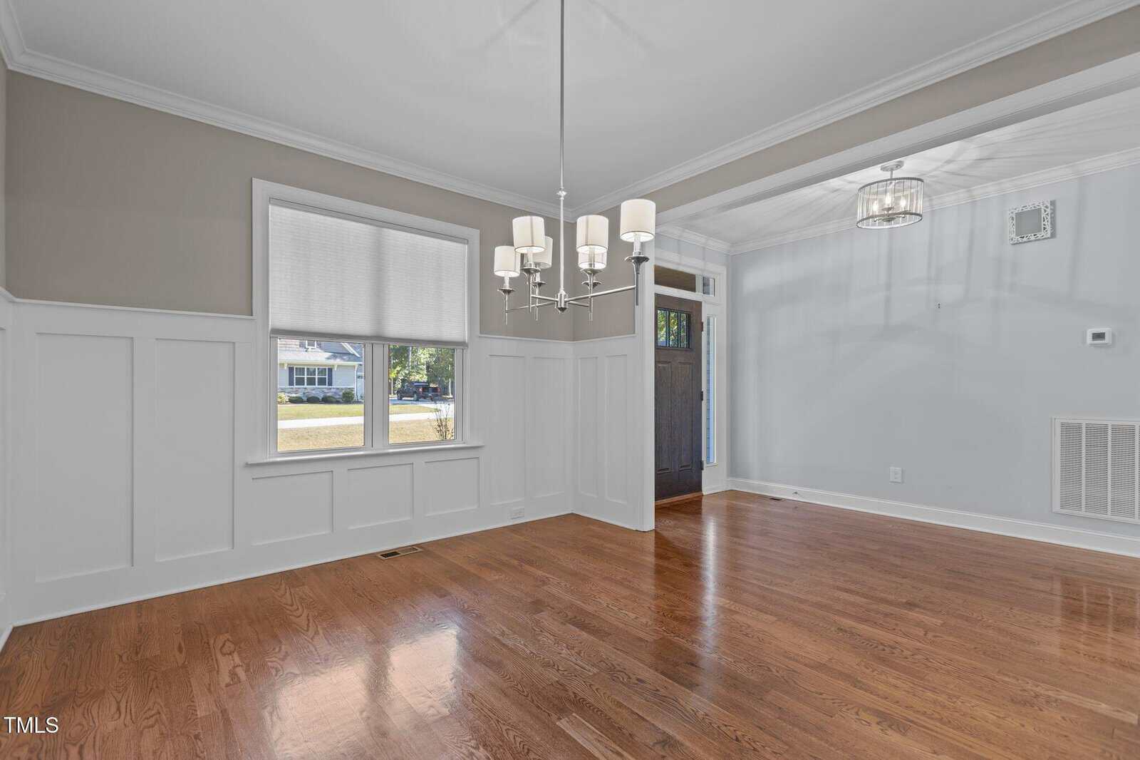 Bright dining room with white wainscoting, tan upper walls, hardwood floors, a large window with a shade, and a modern chandelier. An open entryway leads to another area with a similar light fixture.