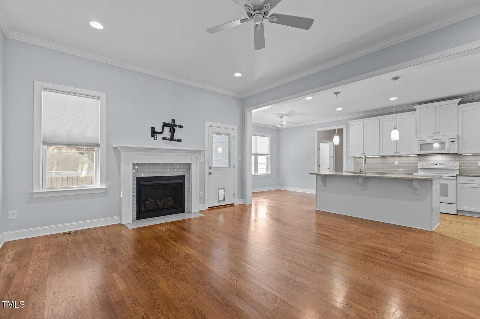 Bright, empty living room with hardwood floors, a white fireplace, and ceiling fan, opening into a modern kitchen with white cabinets, an island, and pendant lights.