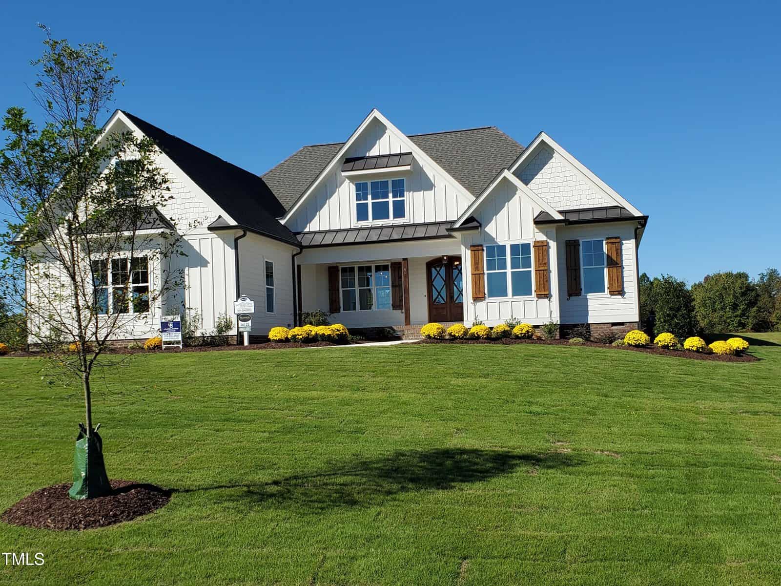 A modern white house with a dark roof, wooden shutters, and a front porch sits on a well-manicured lawn with yellow flowers and a young tree, under a clear blue sky.