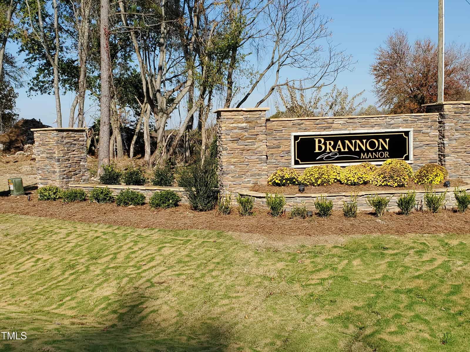 A stone entrance sign reads “Brannon Manor” with landscaping, yellow flowers, and trees in the background on a sunny day.