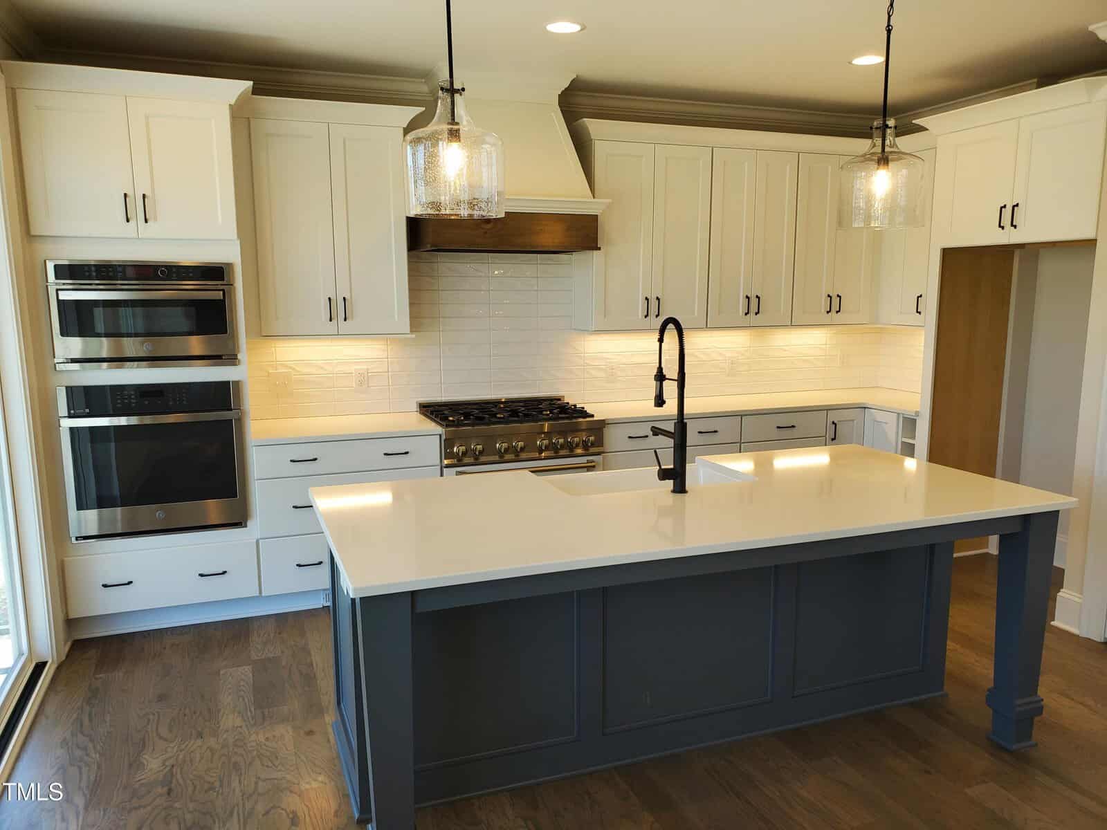 A modern kitchen with white cabinets, a dark center island, stainless steel double oven, large gas stove, farmhouse sink with black faucet, pendant lights, and a white tile backsplash. The floor is dark wood.
