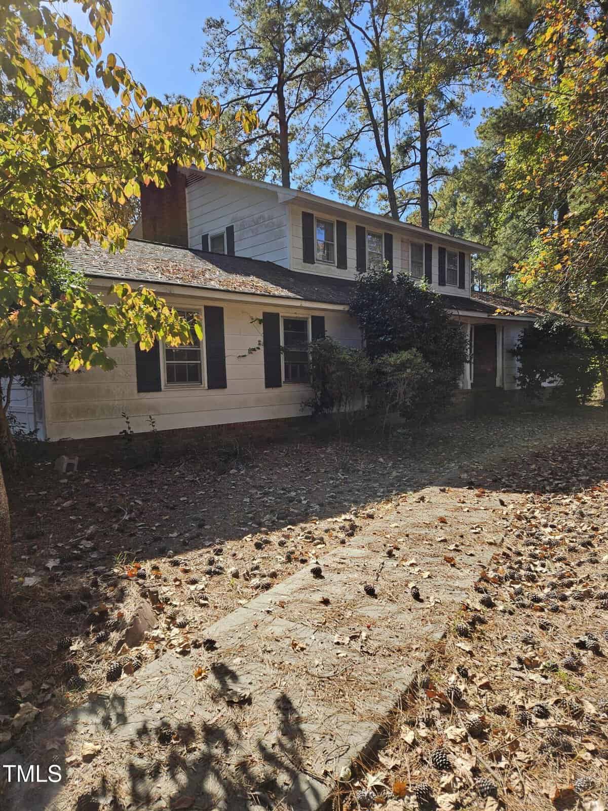 A two-story white house at 109 Dogwood Circle, with a dark roof, is partially shaded by tall trees in Butner. The yard is covered in dry leaves and pine cones, and shrubs line the front of the house. The sky is clear and blue.