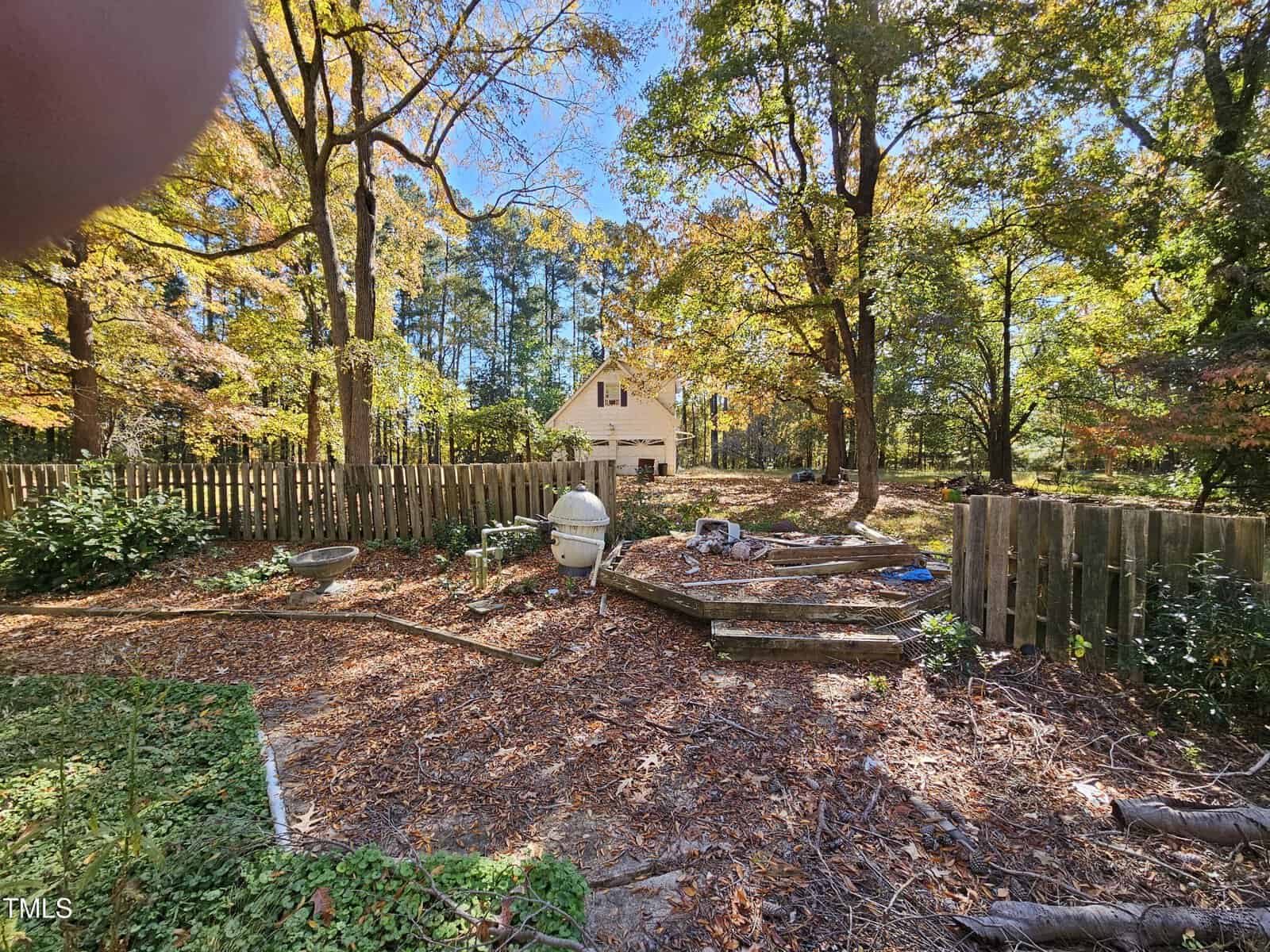 A backyard with a wooden fence, surrounded by trees with autumn foliage. There are raised garden beds and a white structure in the center. A house is visible in the background, partly obscured by trees.