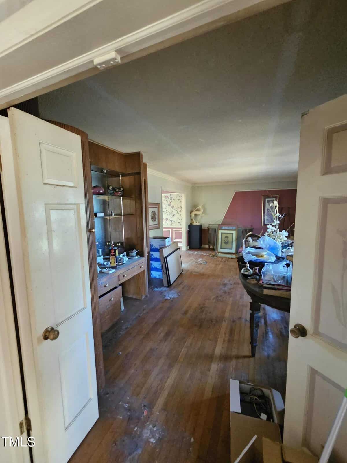 A cluttered room with wooden floors, an open door, and scattered items on a table and floor. Shelves with various objects are on the left, and a framed picture leans against a red wall in the background. Ceiling and floor show signs of wear.