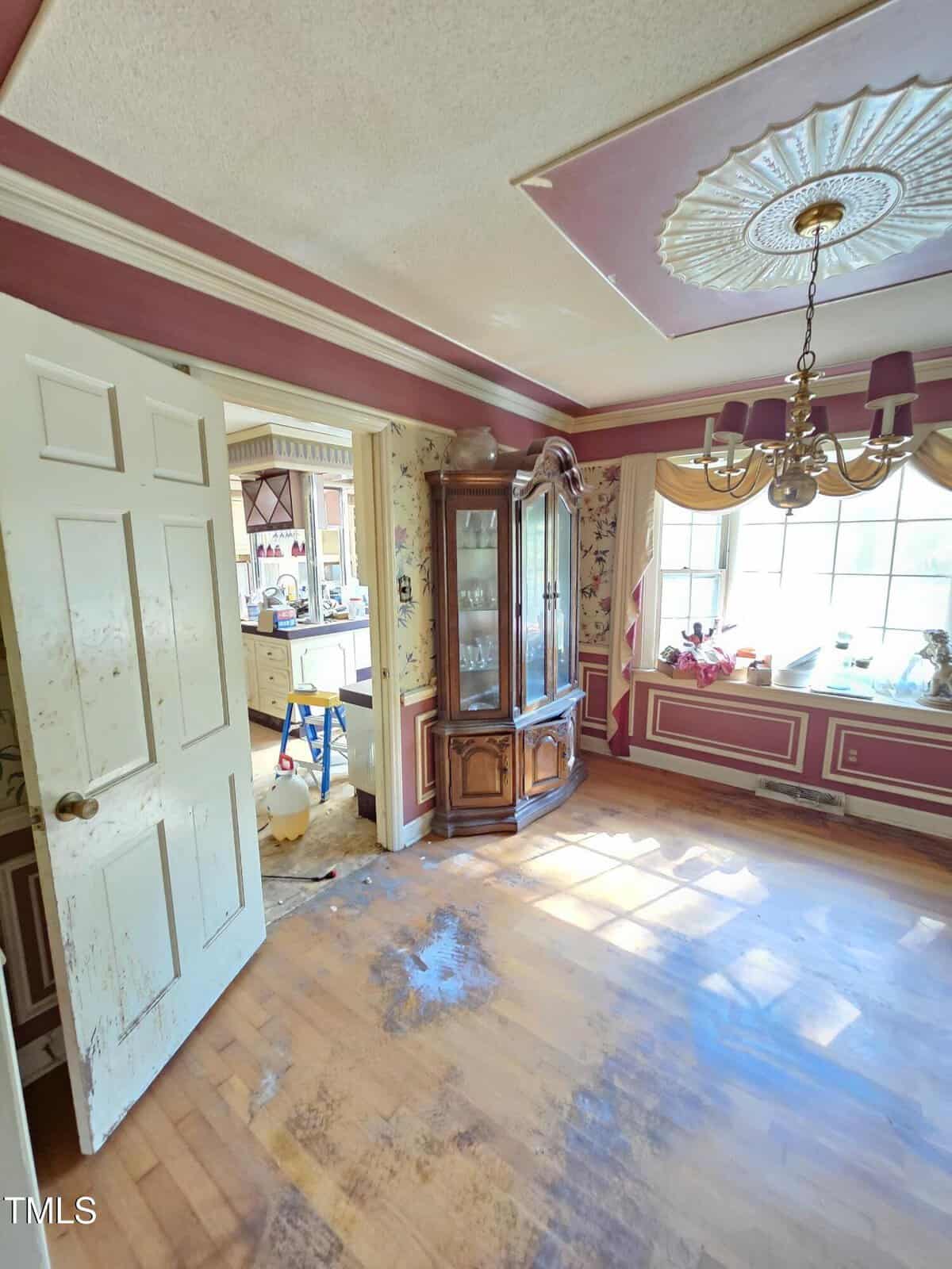 A room with red and beige walls features a ceiling light, decorative molding, and a large window. A wooden cabinet stands in the corner, and a door leads to a kitchen area with visible cabinets and appliances. Sunlight casts shadows on the floor.