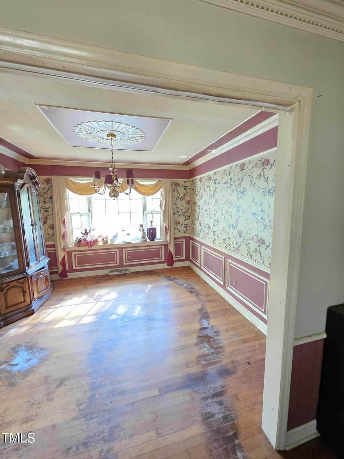A dining room with floral wallpaper, a decorative chandelier, and a large window with yellow drapes. A wooden floor shows signs of wear. A china cabinet is on the left side of the room. Sunlight streams through the window.