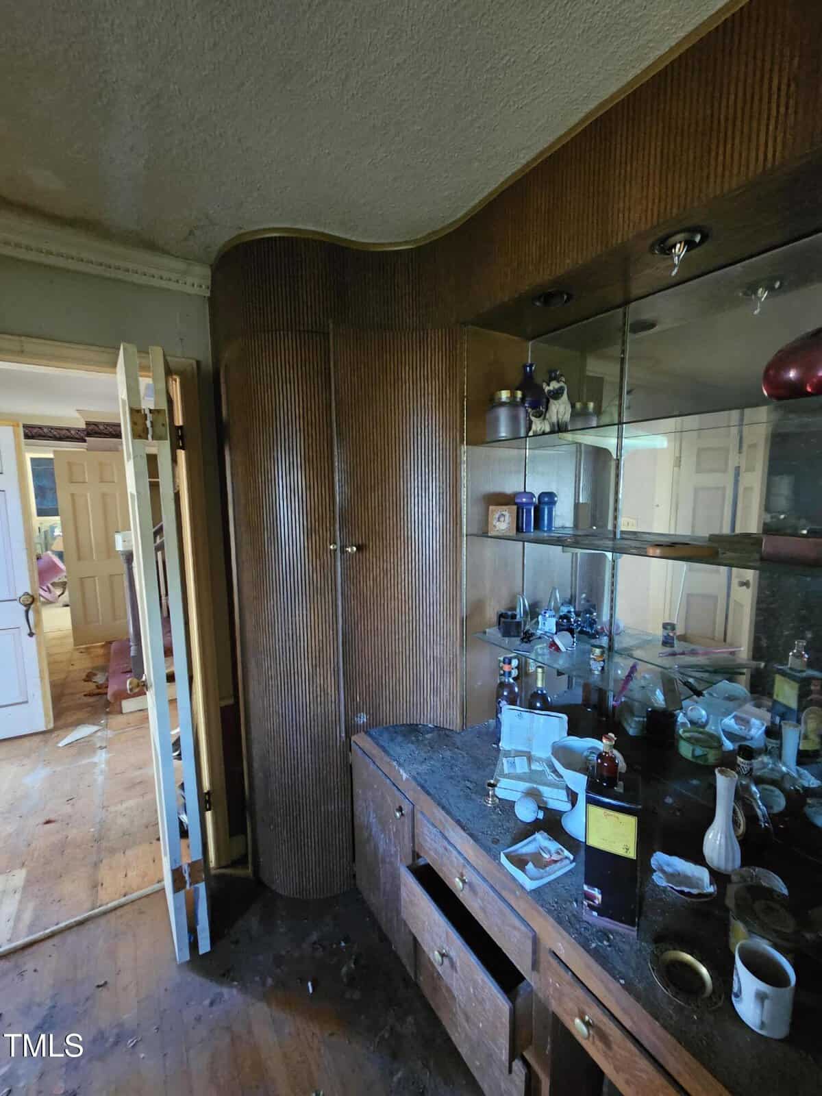 A cluttered room with wooden paneled walls and a mirrored cabinet. The counter has various bottles and miscellaneous items. An open door reveals a messy, partially visible adjacent room. The floor is wooden, littered with debris.