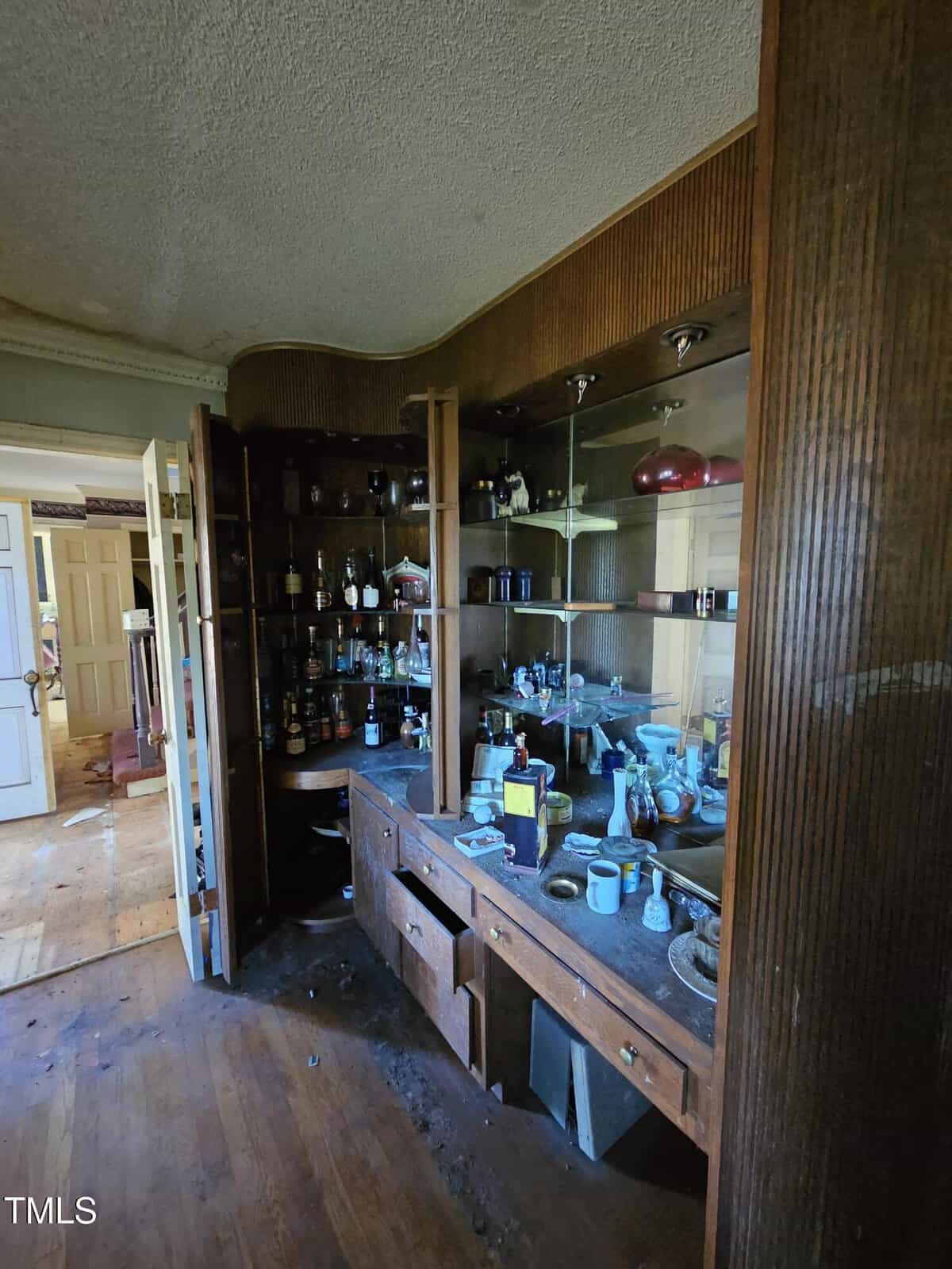A wooden cabinet with glass shelves displaying various bottles and glasses, some cluttered. The room appears dimly lit with a slightly worn wooden floor. An open door on the left leads to another room, revealing more clutter.